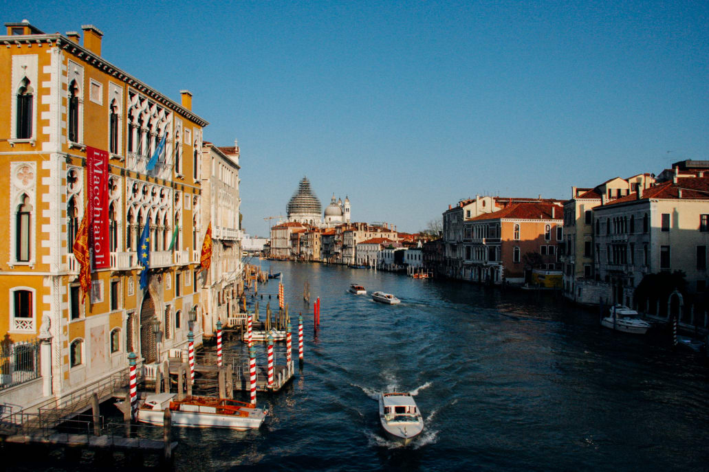 A view of the Grand Canal in Venice with historic buildings lining the sides, boats on the water, and a distant dome under restoration scaffolding.