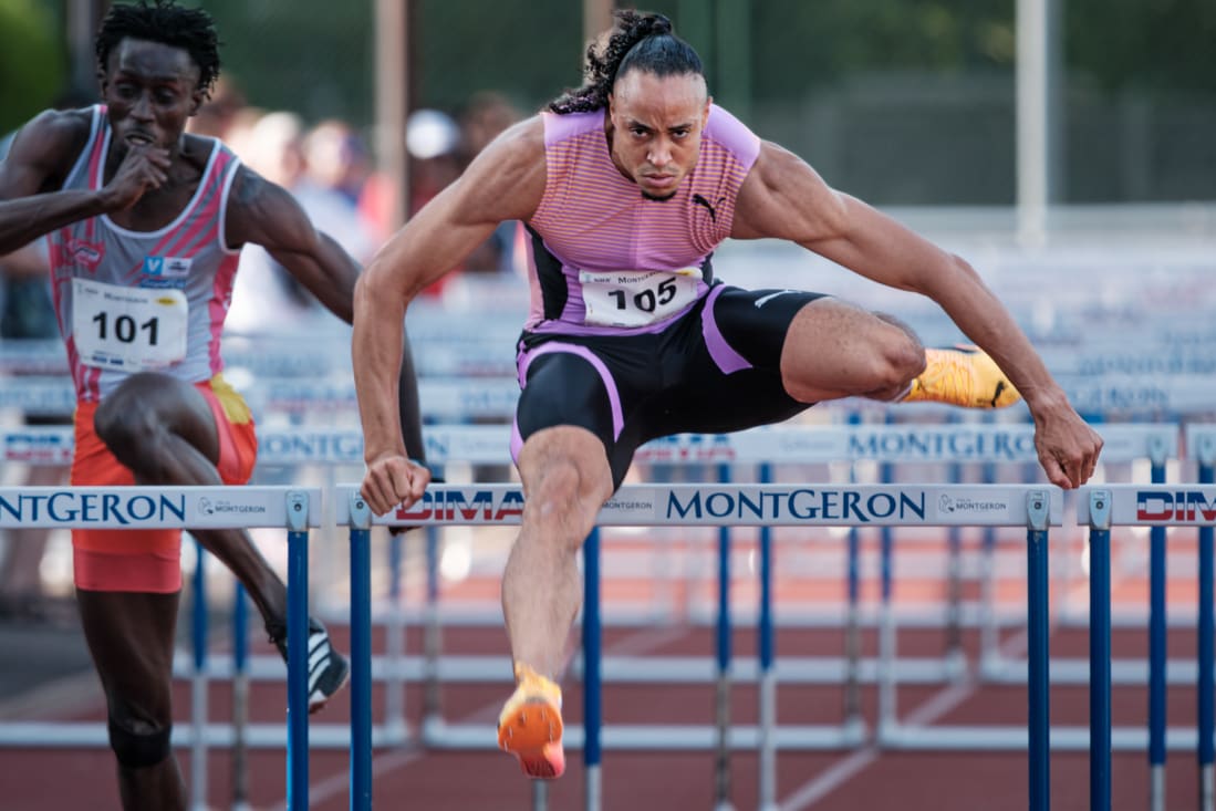 A male athlete in a purple and pink uniform is leaping over a hurdle during a race. A second athlete is also in motion behind him.