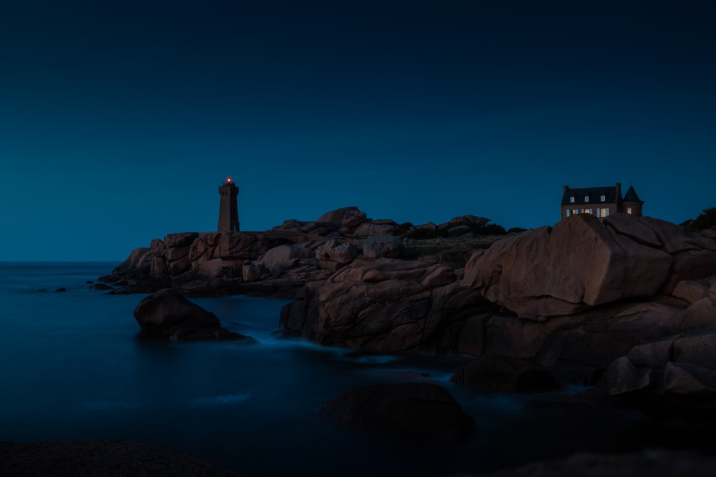 A lighthouse with a shining beacon on a rocky coastline at dusk, with a house nearby and calm ocean waters in the foreground.