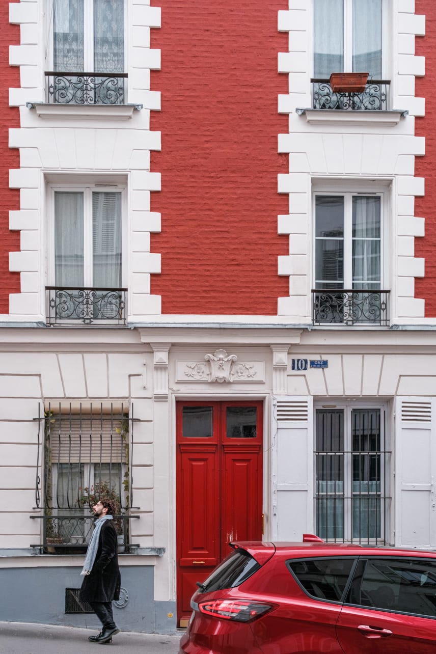 A man walks past an old red-painted brick building with white window frames and a red door. A red car is parked in the foreground.
