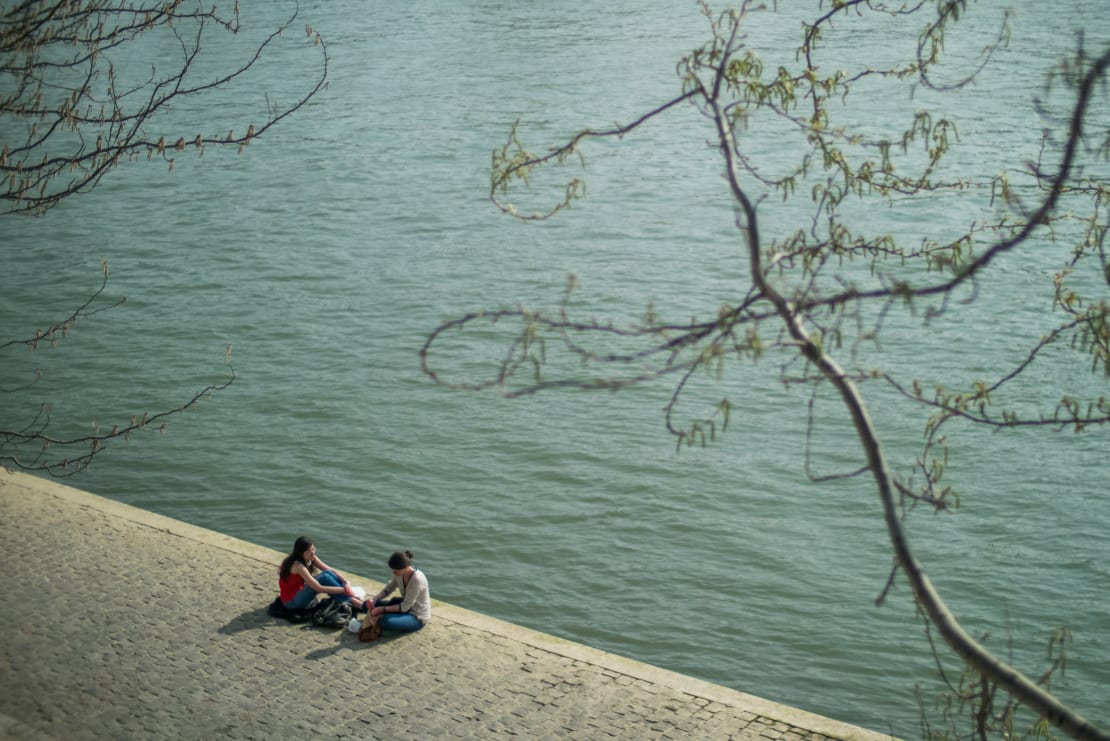 Two people sitting by a body of water with a tree branch in the foreground.