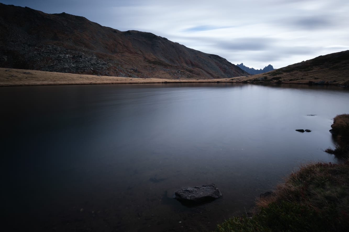 A serene landscape featuring a smooth lake with a single rock near the shore, surrounded by rolling hills under a cloudy sky.