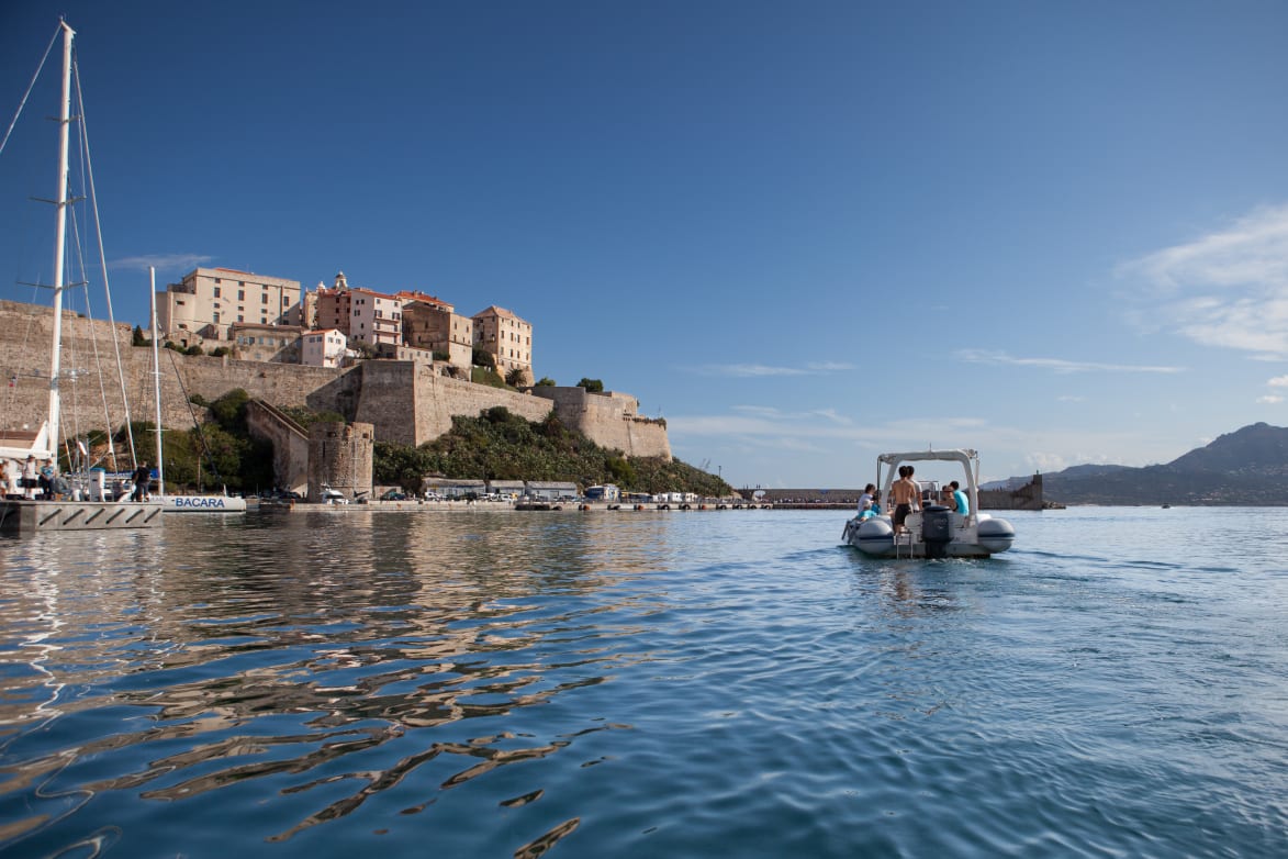 A small boat is cruising through calm waters near a coastal town with historical buildings and a fortress on the hillside.