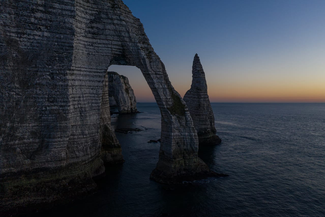 A coastal landscape featuring striking white cliffs and rock formations, including a large arch and a pointed spire, set against a tranquil sea and twilight sky.