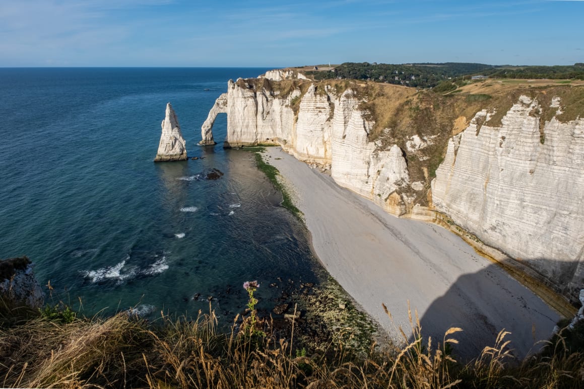 A coastal landscape featuring dramatic white chalk cliffs with an arch formation, surrounded by the blue sea and a beach.