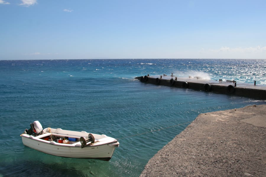 A small white boat anchored near a concrete pier with calm, clear blue ocean water and a cloudless sky.