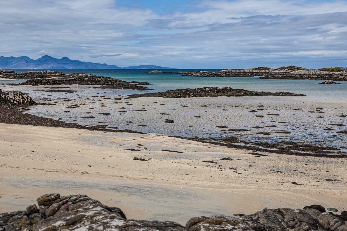 Photo of a white sandy beach, with turquoise sea, and in the background low mountains