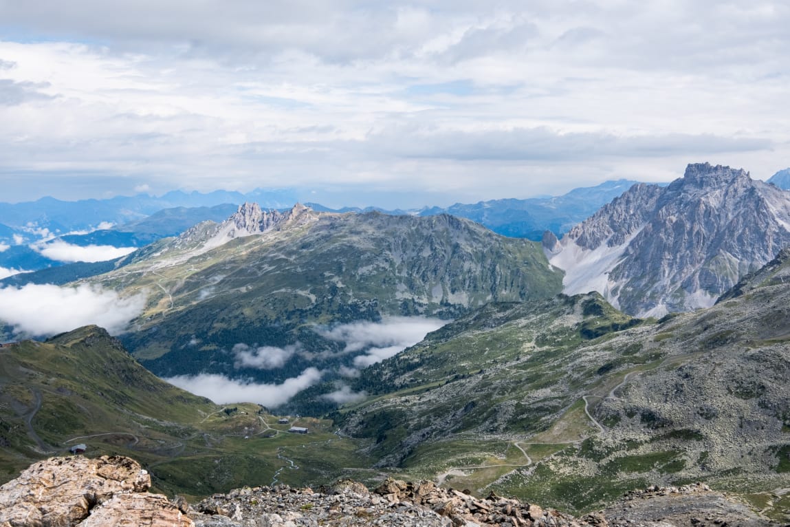 A panoramic view of a mountainous landscape featuring lush green hills, rocky peaks, and patches of clouds in the valleys.