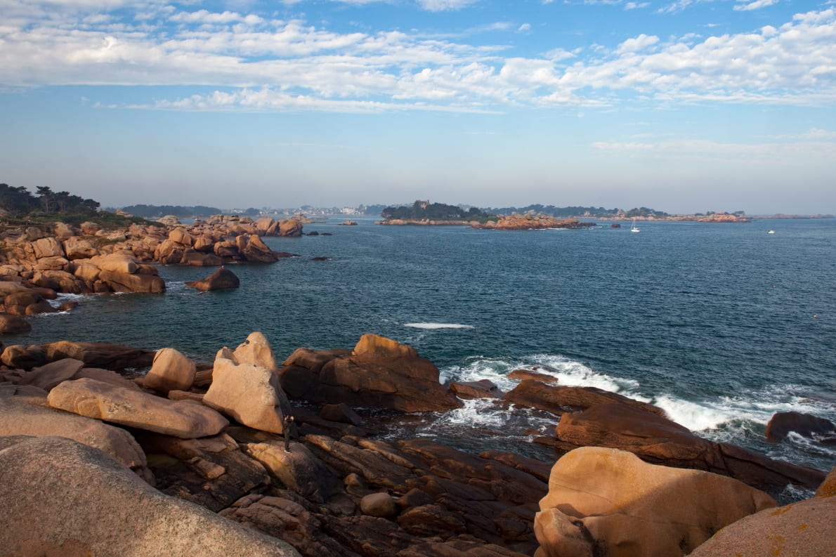 A coastal scene featuring large, weathered rocks along the shoreline, with gentle waves lapping at the base. In the distance, an island with some greenery and a hint of buildings is visible under a cloudy blue sky.