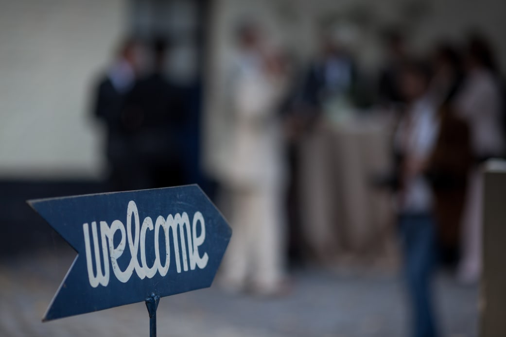 A "welcome" sign in the foreground with a blurry background showing a group of people.