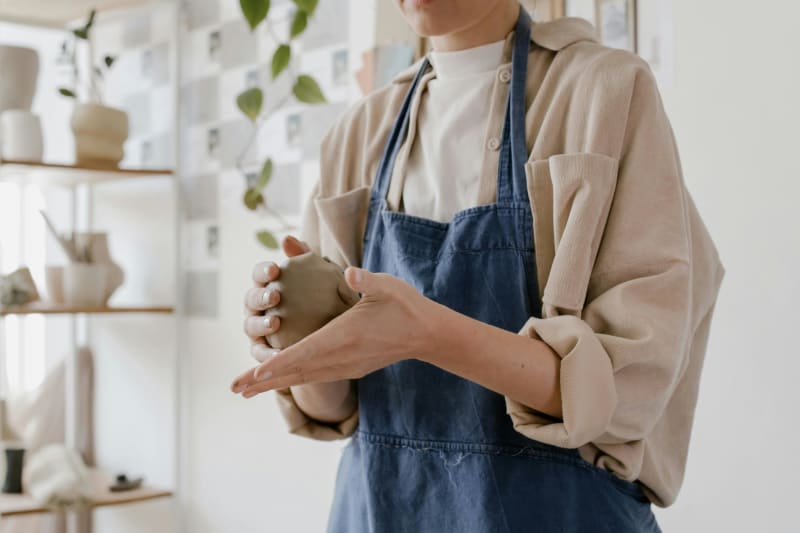 A ceramist in a denim apron holding a clay vessel in progress, shelves of finished pottery behind them