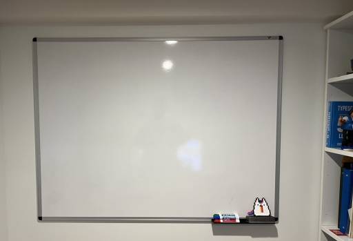 My office's back wall has a whiteboard on it. There are erasable markers, an eraser, and a small flat plastic cute cat throwing up a rainbow. To the right is my bookshelf.