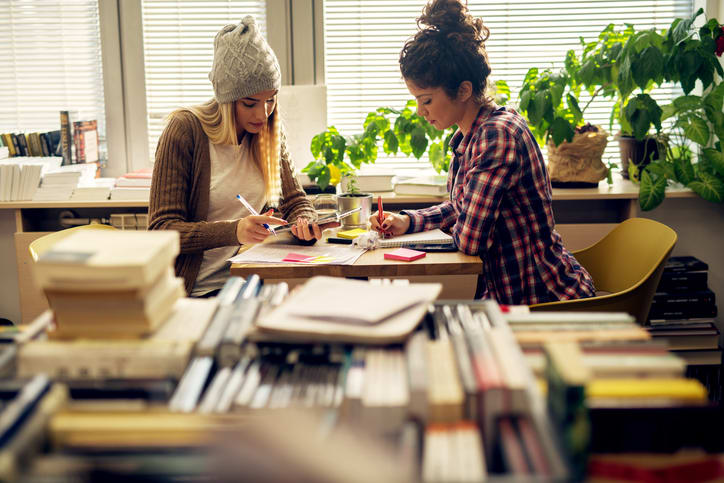 Hipster student female friends sitting in the sunny school library near the window and doing homework together with notes and a tablet.