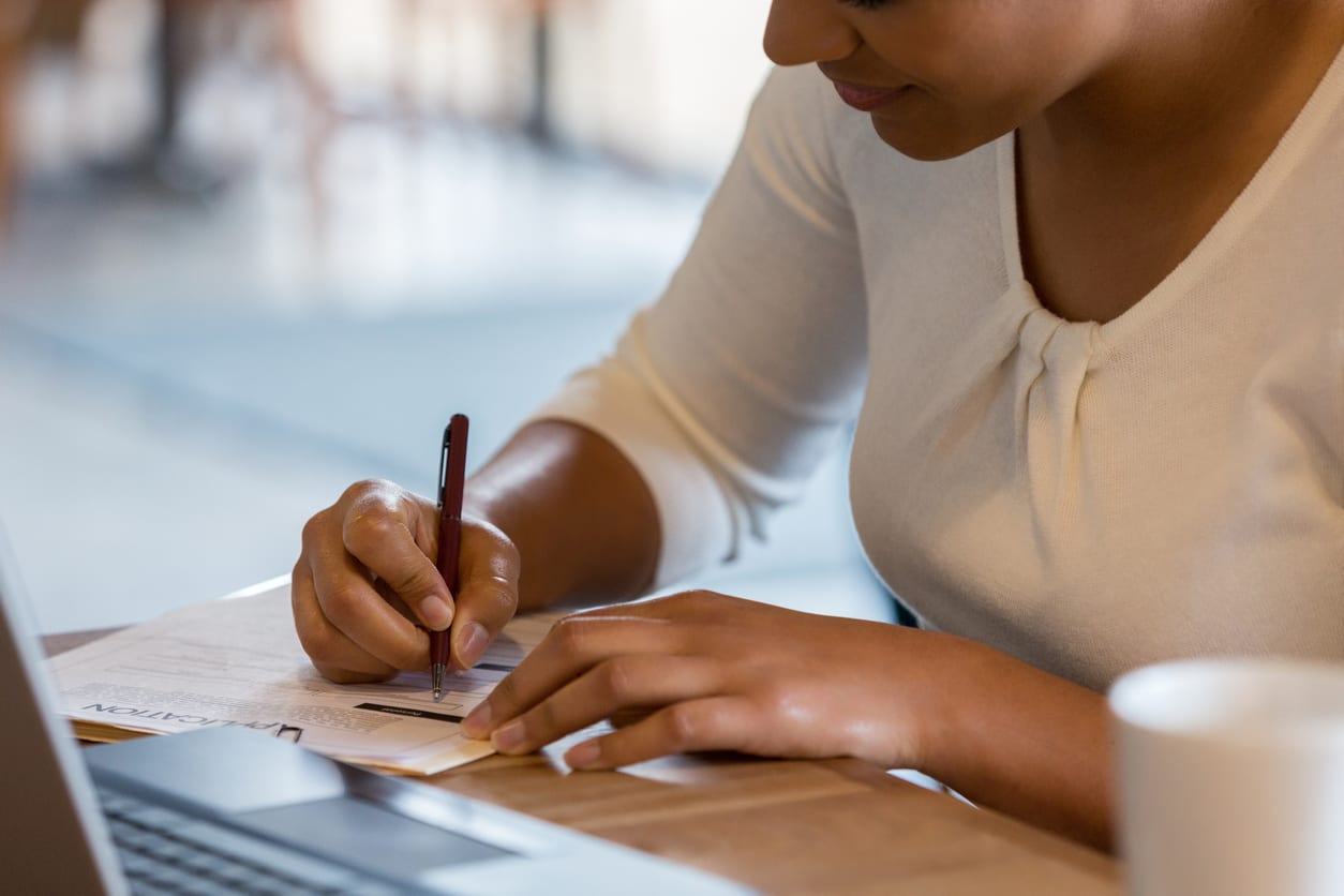 An unrecognizable Hispanic woman references the information on her laptop to fill out a job application at the coffee shop.