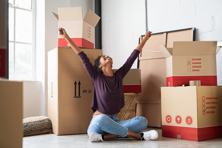 Happy young woman sitting in new apartment and raising arms in joy after moving in. Joyful and excited african girl moving to new home. Black woman sitting on floor in her house.