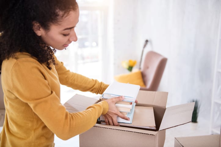 Necessary books. Charming curly-haired girl fitting a pile of books into the box while preparing to move out of an old flat