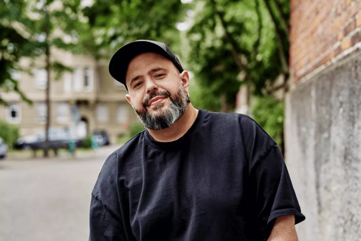 Foto von Enrico Kunze mit Basecap und schwarzem T-Shirt am Straßenrand vor Mehrfamilienhäusern