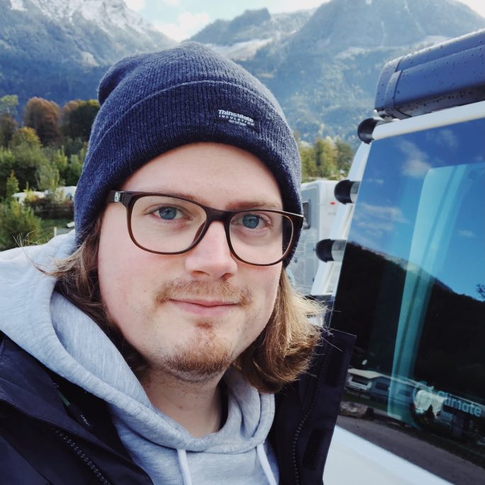 nion Mitarbeiter Martin mit Brille, Mütze und Kapuzenpulli blickt lächelnd in die Kamera, im Hintergrund Berge