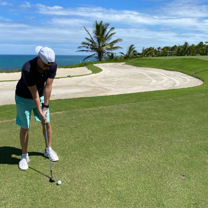 Sebastian in Shorts und T-Shirt hält einen Golfschläger, bereit den Ball zu Schlagen, im Hintergrund Palmen und das Meer