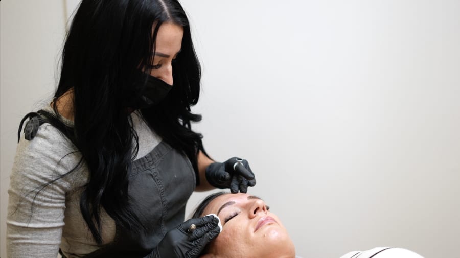 Sarah performing microblading strokes on a reclined client, gloved and masked, inside her Nirvana PMU studio
