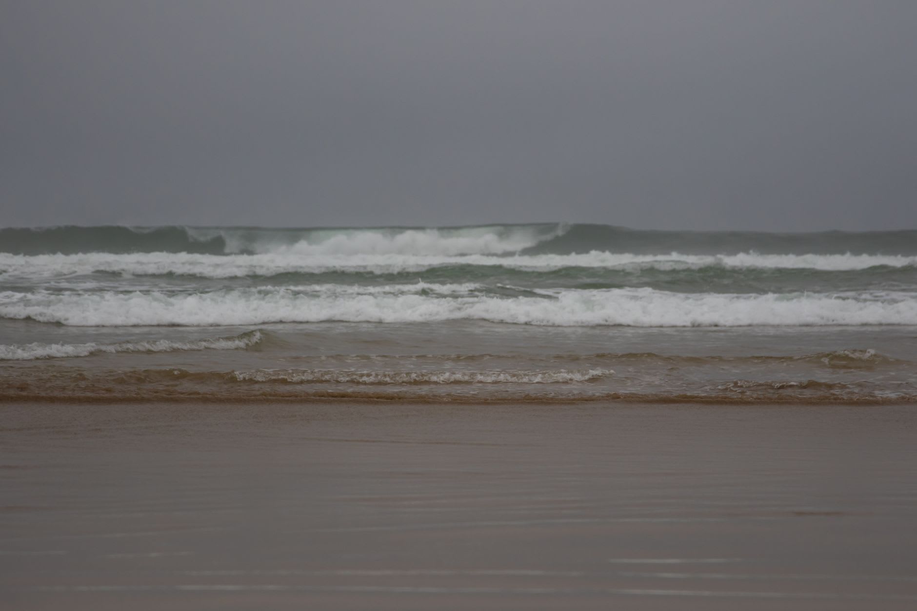 Vagues qui déferlent sur une plage de sable brun sous un ciel gris et bas, mer agitée et écume