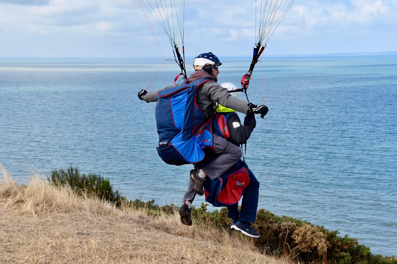 Deux parapentistes en biplace courent sur une pente herbeuse face à la mer au moment du décollage