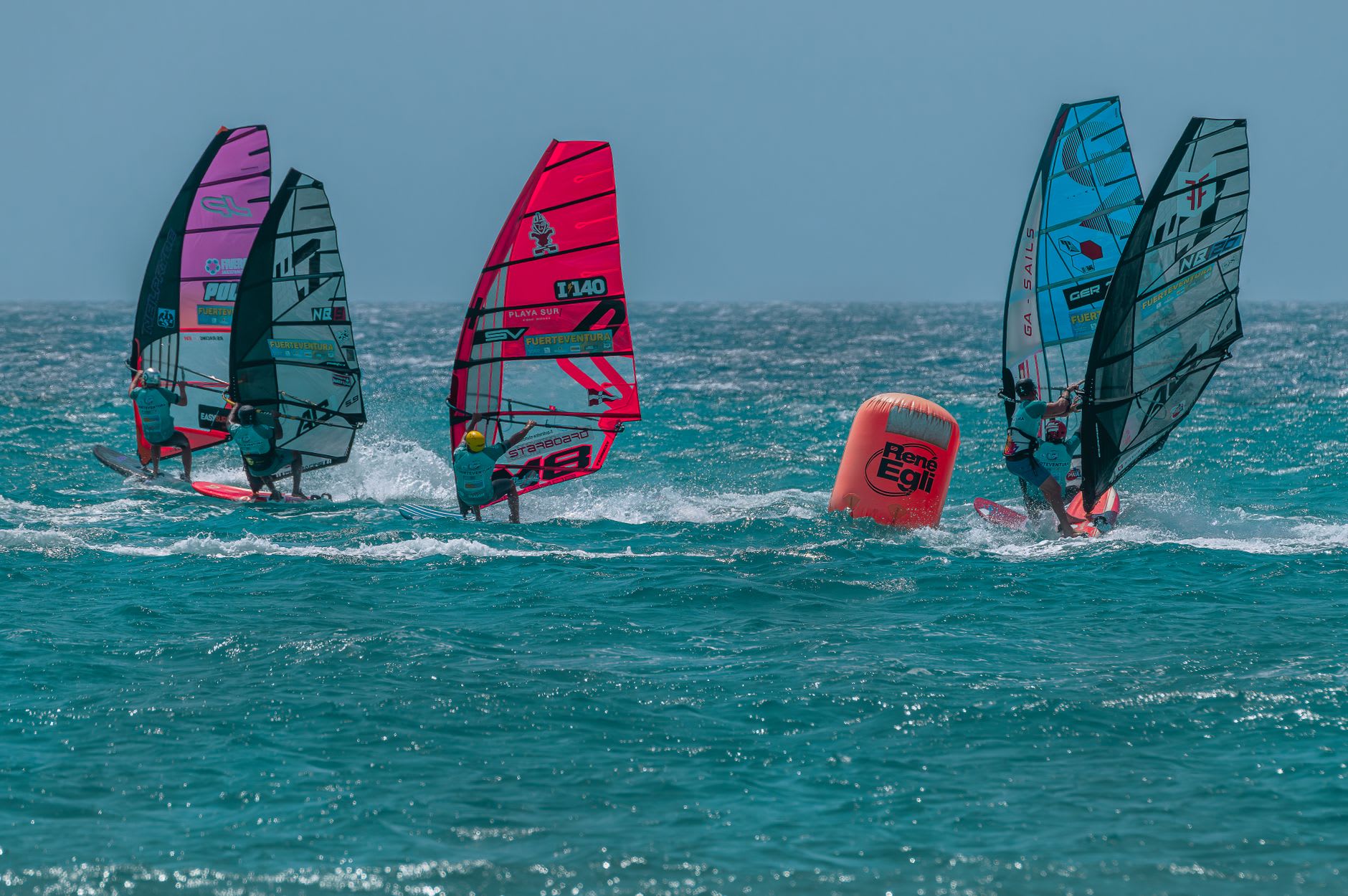 Deux planches à voile filant au près sur une mer moutonneuse, voiles rouge et rose tendues par un vent soutenu, moutons blancs jusqu’à l’horizon