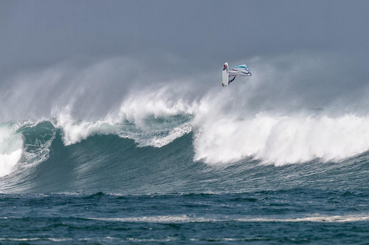 Planchiste à voile en action sur la houle, plusieurs ailes de kitesurf gonflées dans le ciel en arrière-plan, mer turquoise agitée