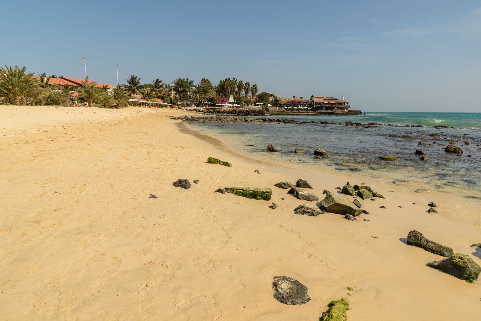 Plage de sable doré parsemée de roches volcaniques noires, rangée de cocotiers et bâtiments à toit rouge en arrière-plan, mer turquoise sous un ciel dégagé