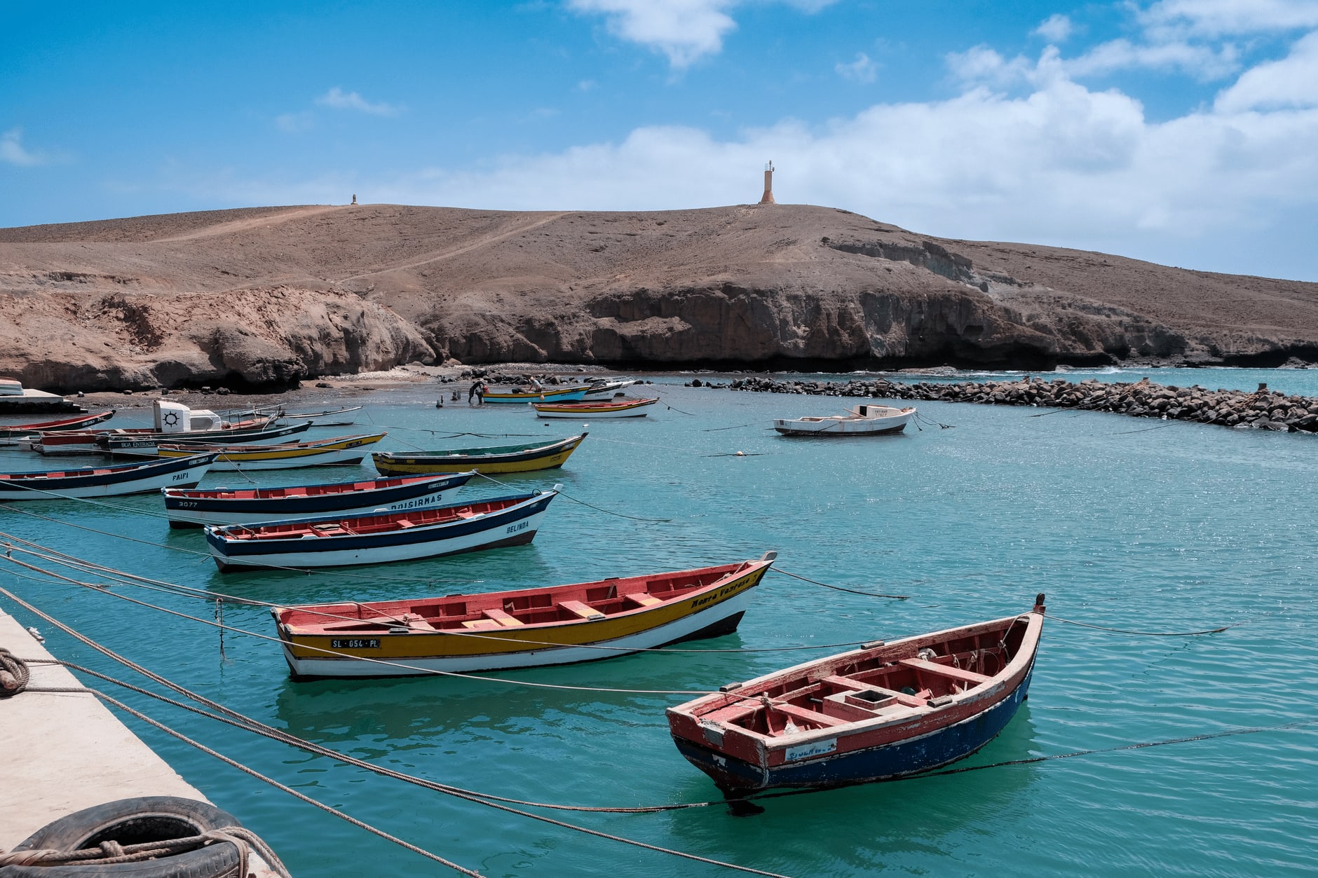Barques de pêche en bois rouge, jaune et bleu amarrées dans le port abrité de Pedra de Lume, sur Sal, derrière une digue de roches noires, colline pelée et petit phare au sommet