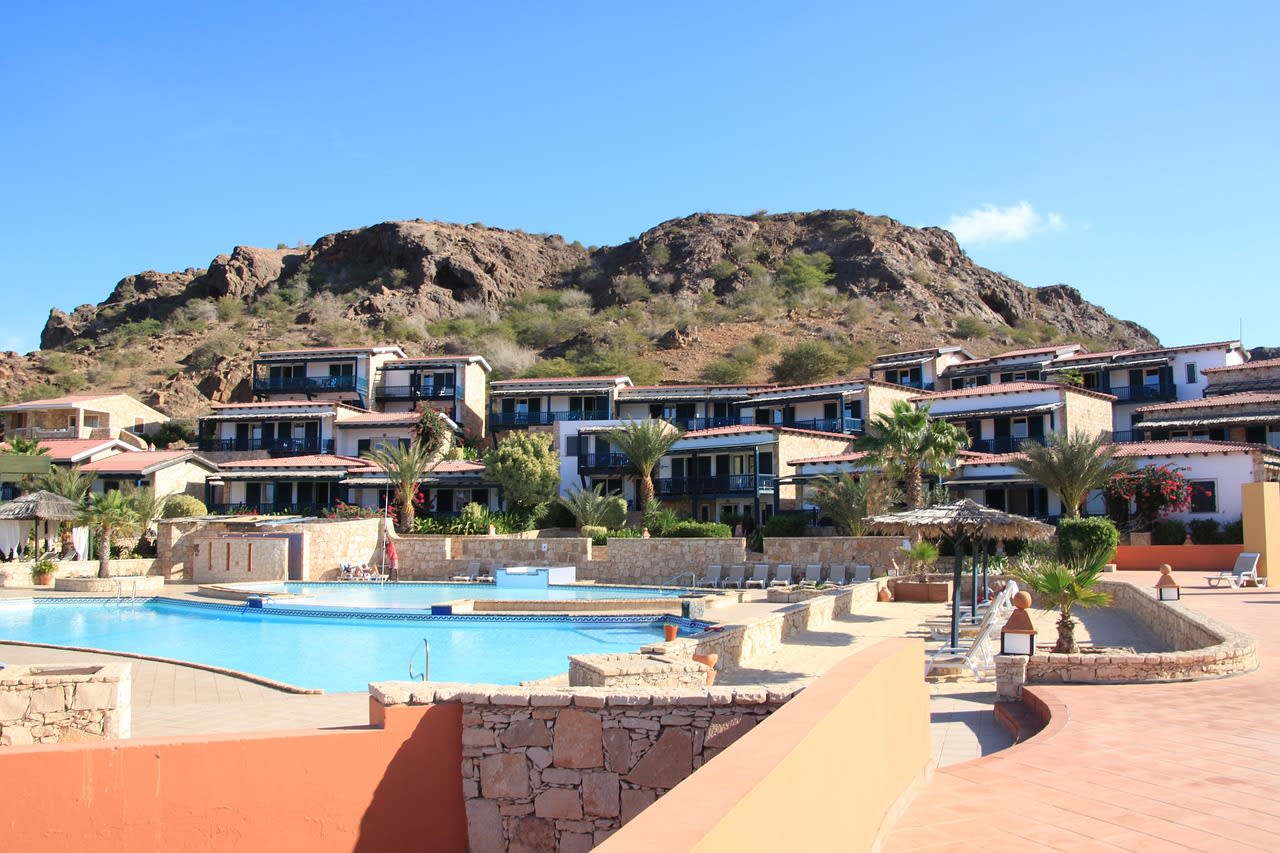 Piscine d’un complexe hôtelier bordée de transats et de petits palmiers, immeubles en pierre à toit de tuiles adossés à une colline volcanique aride, ciel bleu vif