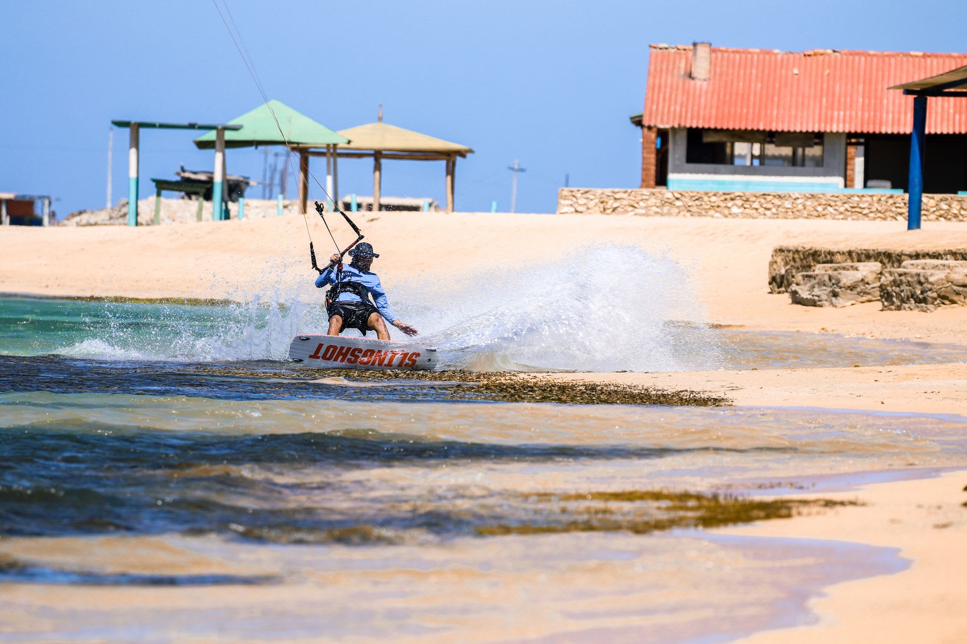 Rider tracté par son aile au ras du bord, gerbe d’eau soulevée, plage de sable ocre et cabanons sous un ciel bleu