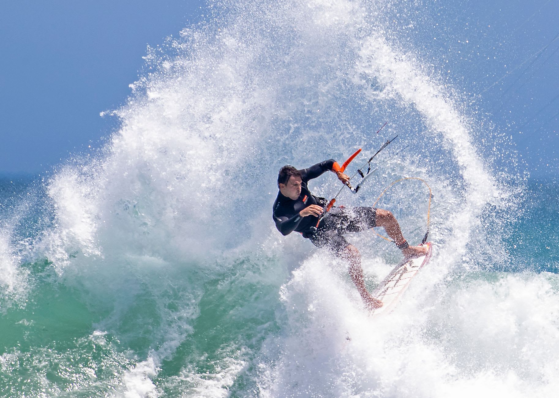 Kitesurfeur sculptant un mur d’écume dans une vague, projections d’eau tout autour sous un ciel bleu franc