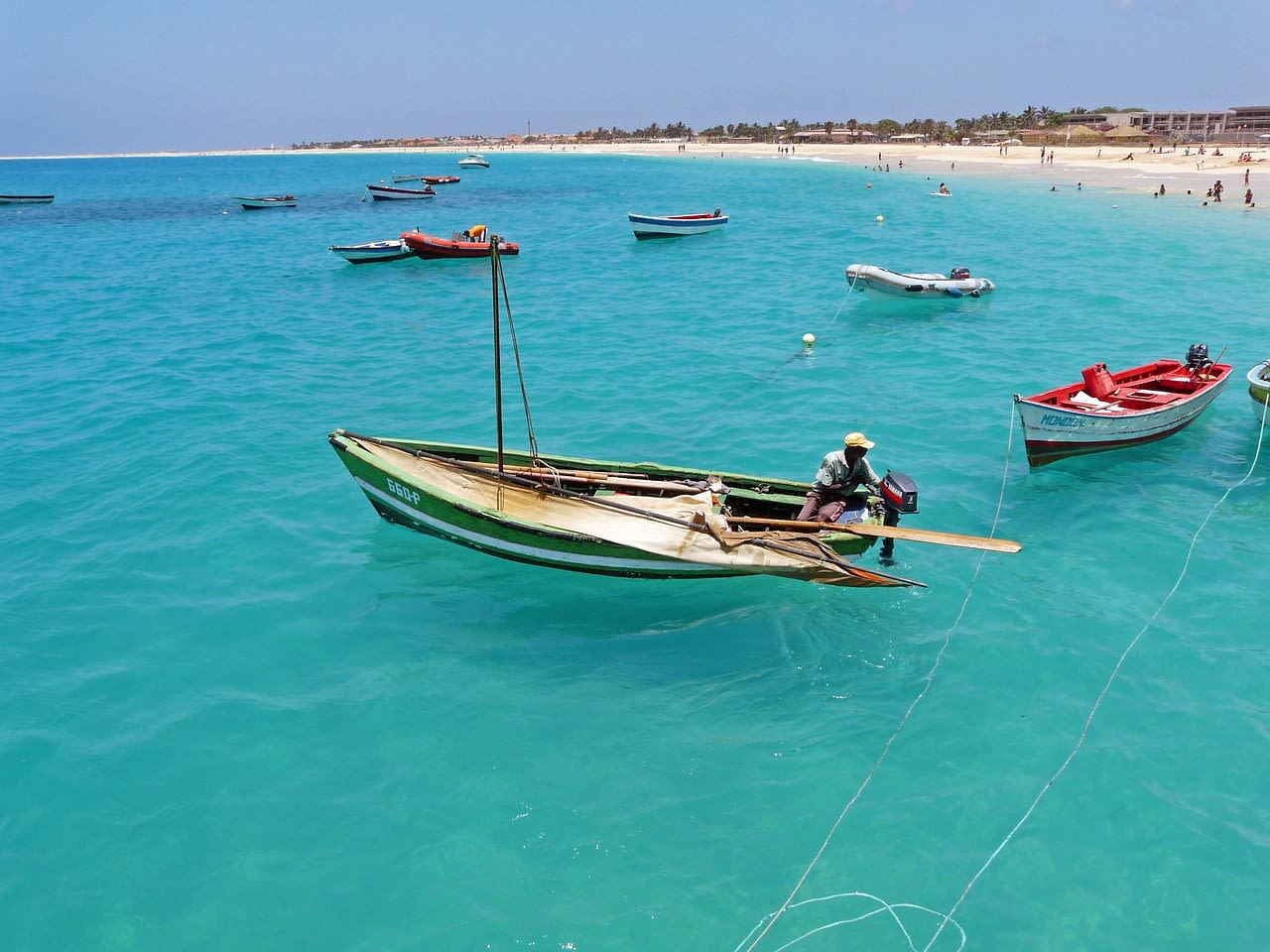 Barques de pêche colorées immobiles sur une eau turquoise et plate, plage et village clair au fond, mer sans vagues