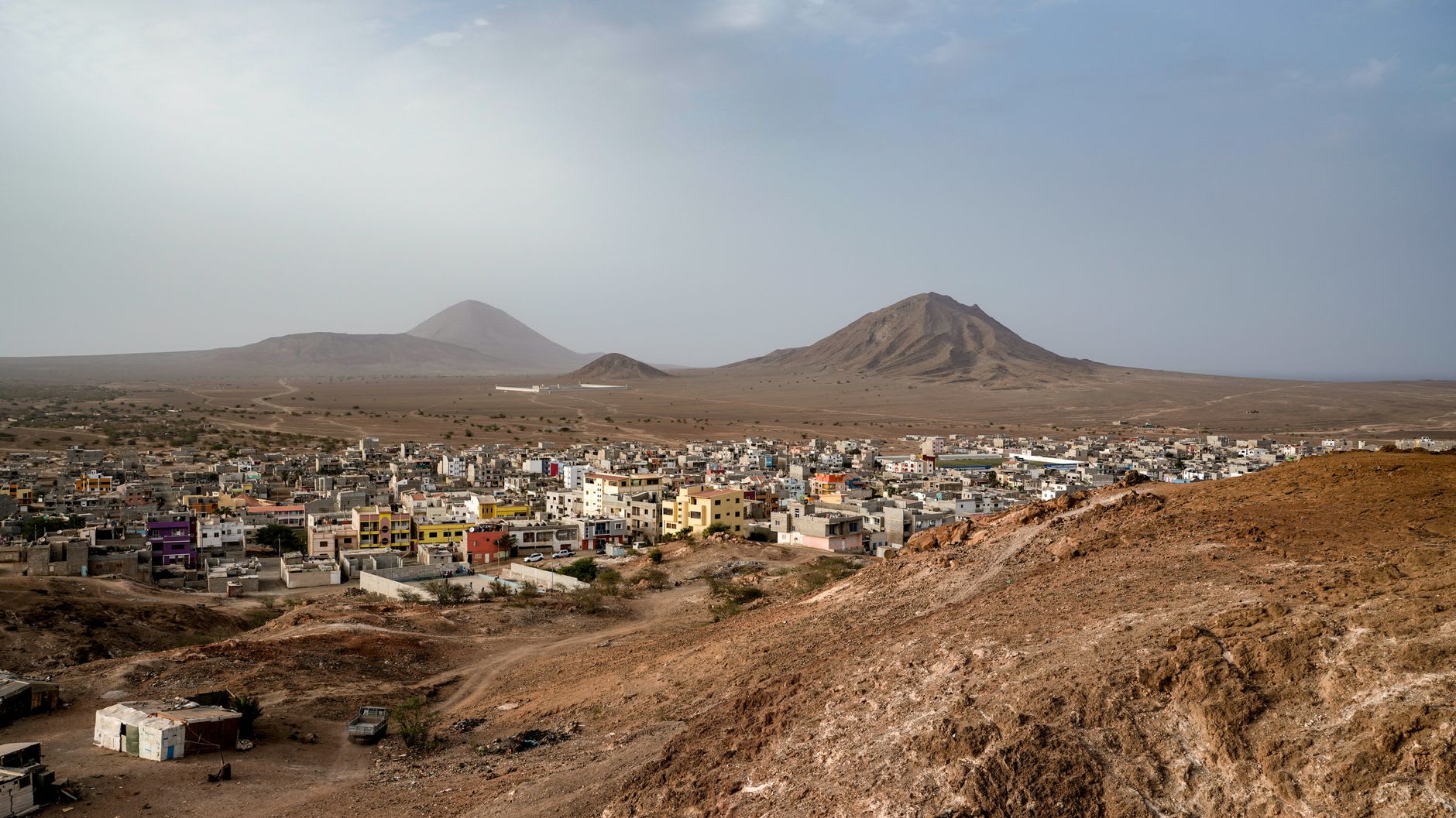 Bourg aux maisons colorées étalé dans une plaine sèche, deux cônes volcaniques à l’horizon sous un ciel voilé de poussière