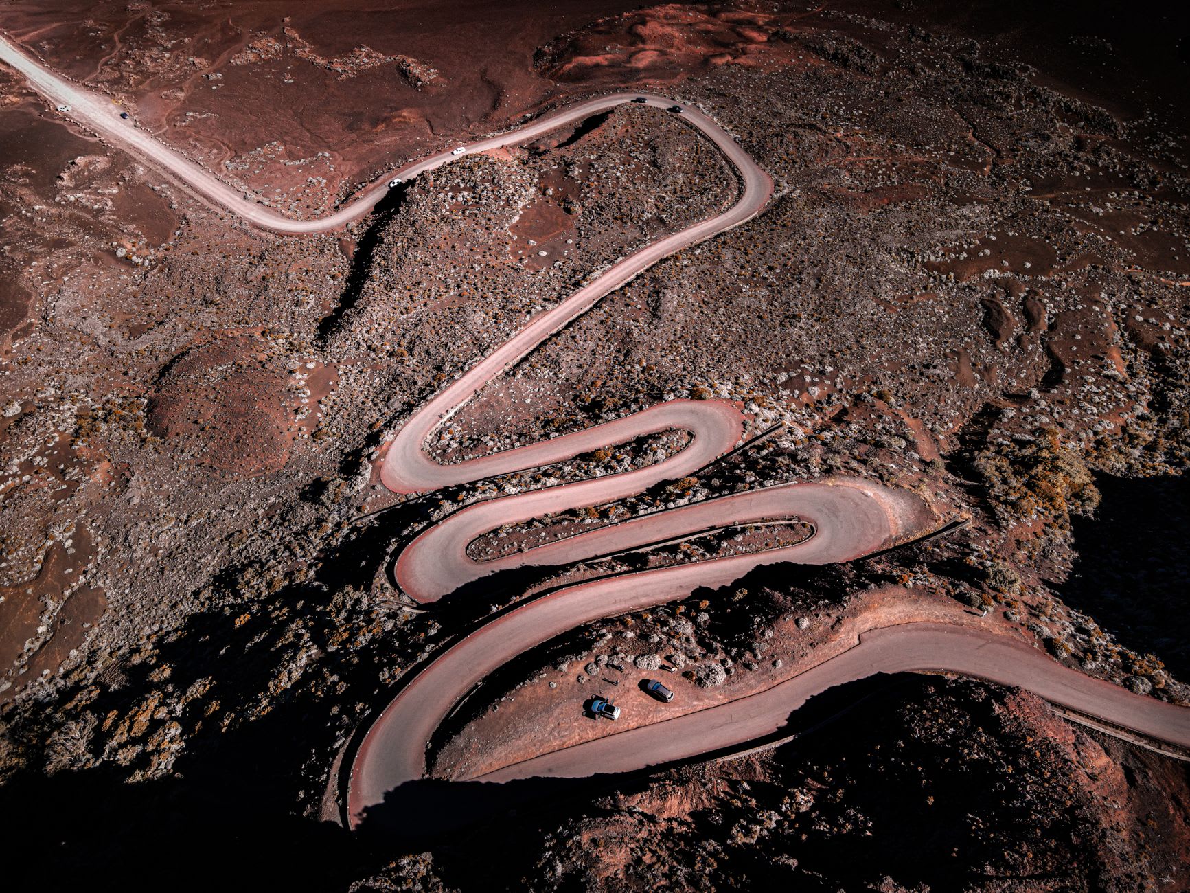 Route en lacets serrés traversant un plateau volcanique rouge sombre, quelques voitures à l’arrêt sur le bas-côté