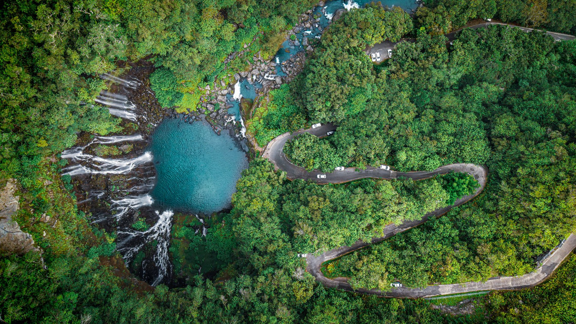 Vue aérienne d’une route de montagne en épingle longeant une cascade et un bassin turquoise en pleine forêt