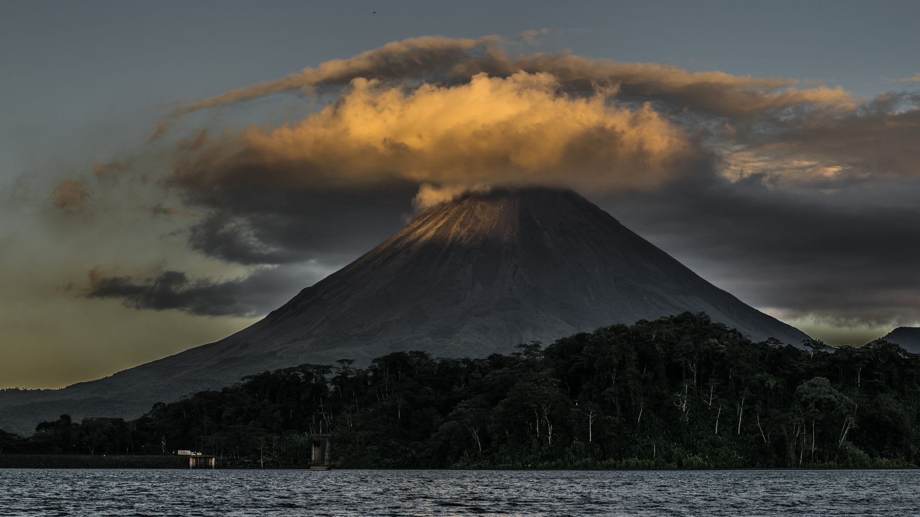 Cône du volcan Arenal coiffé d’un chapeau de nuages orangés au crépuscule, au-dessus du lac