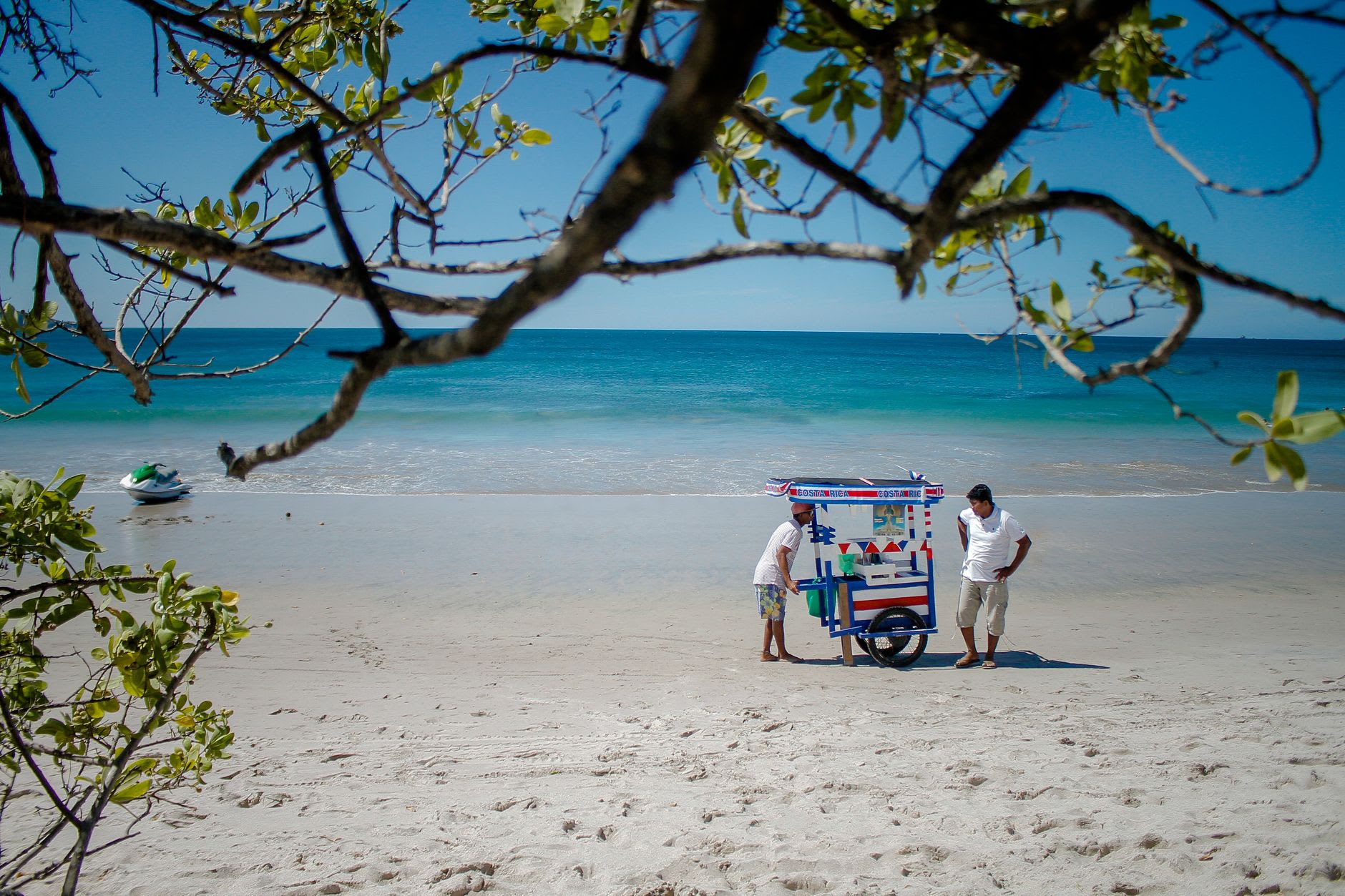 Charrette de vendeur de glaces pilées sur une plage de sable blanc bordée d’eau turquoise, sous les branches d’un arbre