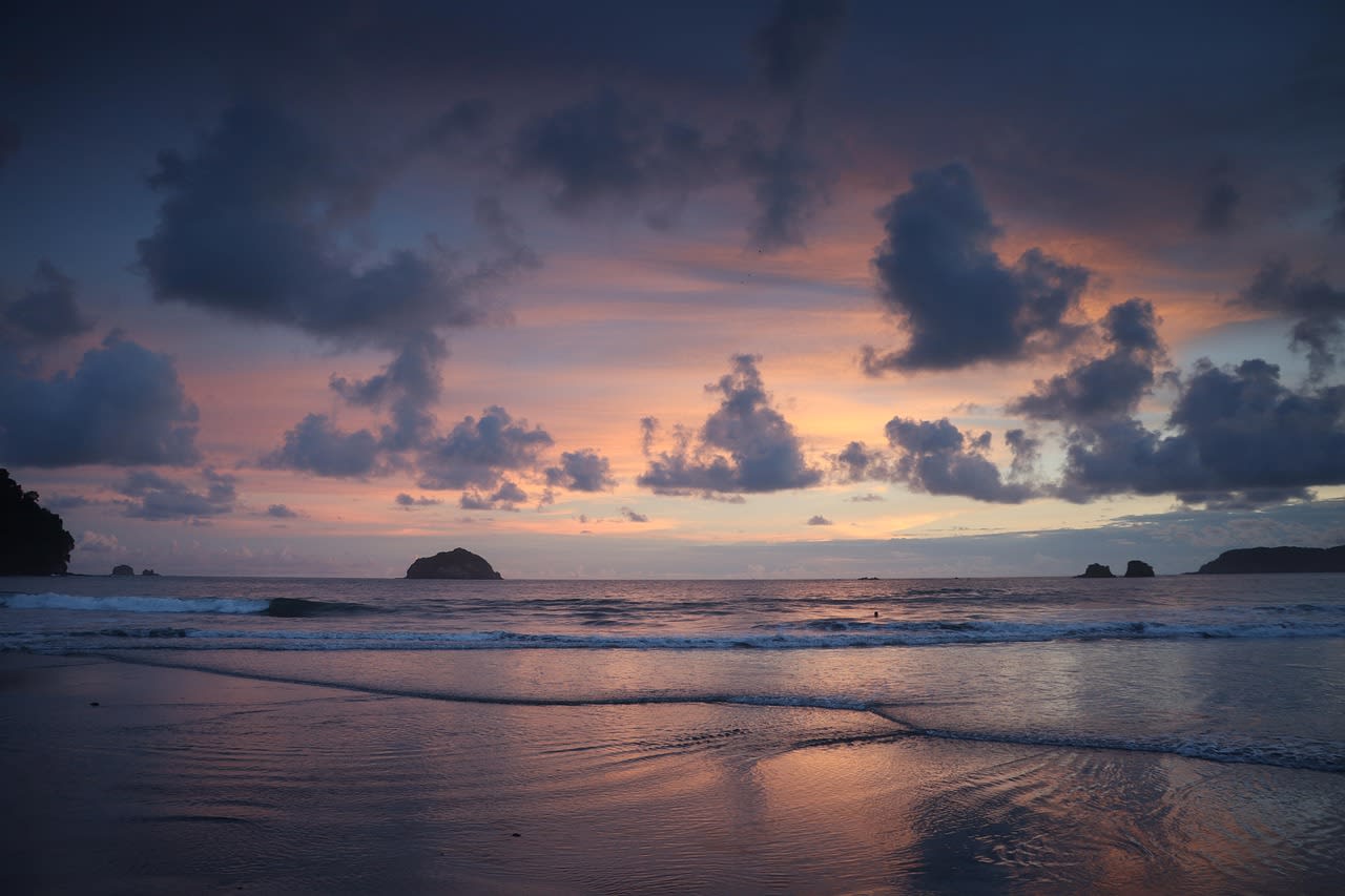 Ciel de fin de journée violacé et nuageux au-dessus d’une plage humide et vide, îlots rocheux au large