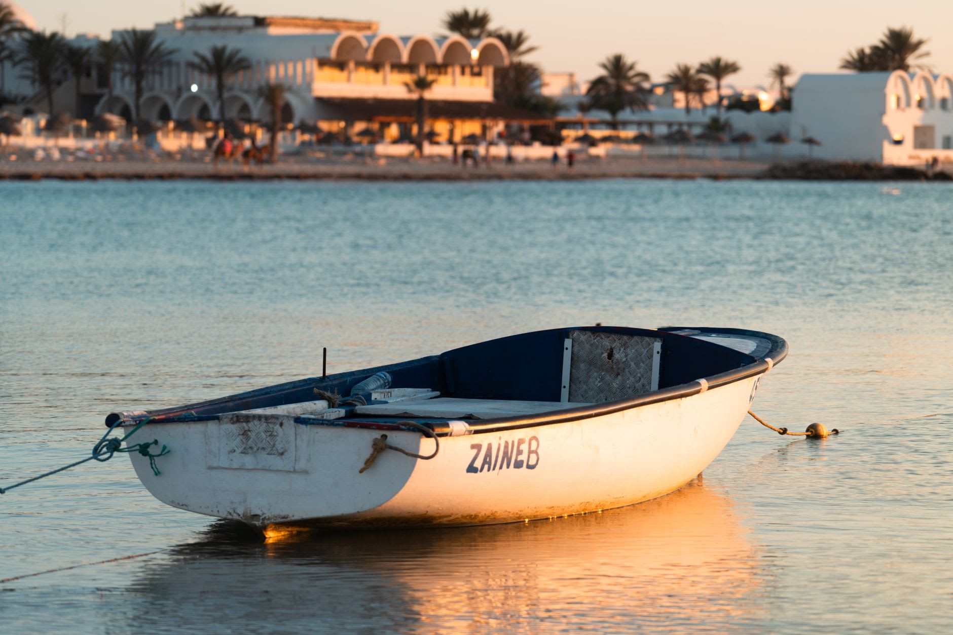 Petite barque de pêche blanche posée sur une eau plate et sans vague, hôtel de bord de plage et palmiers dans la lumière du couchant