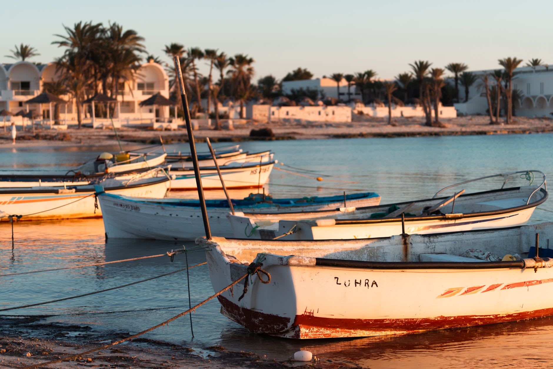 Barques de pêche blanches amarrées dans le lagon calme de Houmt Souk au coucher du soleil, maisons à coupoles et palmiers en arrière-plan