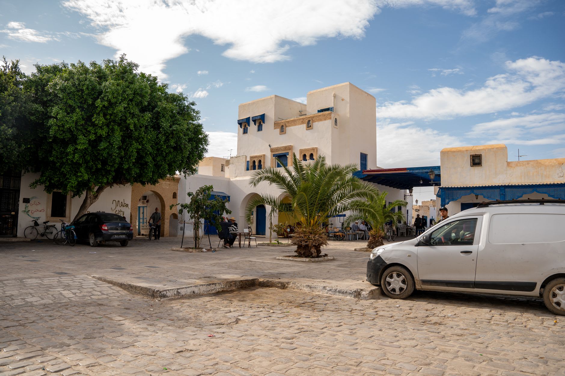 Place pavée d’un village de l’intérieur de Djerba, maisons blanches à volets bleus, grand arbre et terrasse de café en plein jour