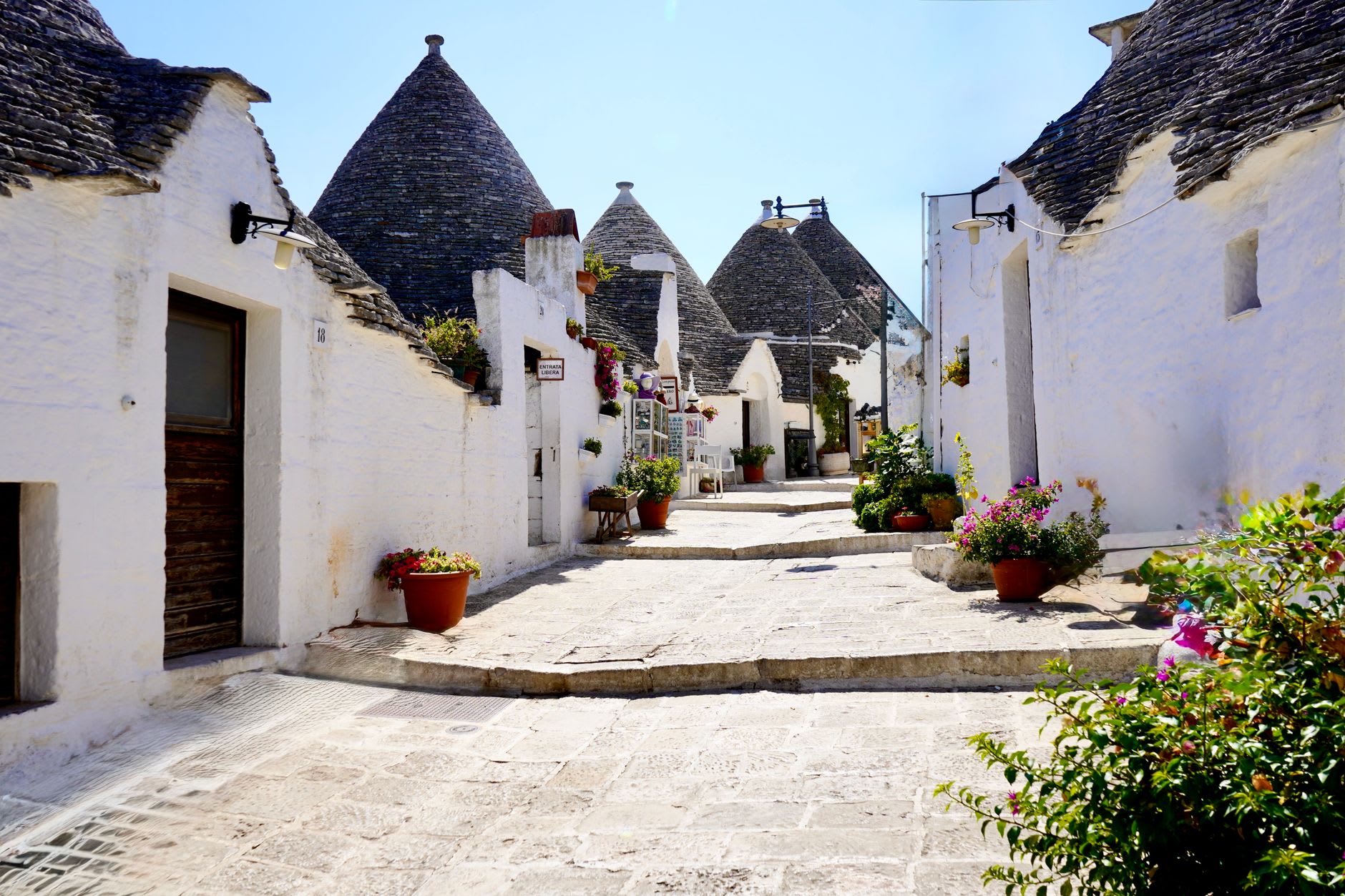 Ruelle pavée bordée de maisons blanches à toit conique, présentoirs de souvenirs et panneau Entrata libera