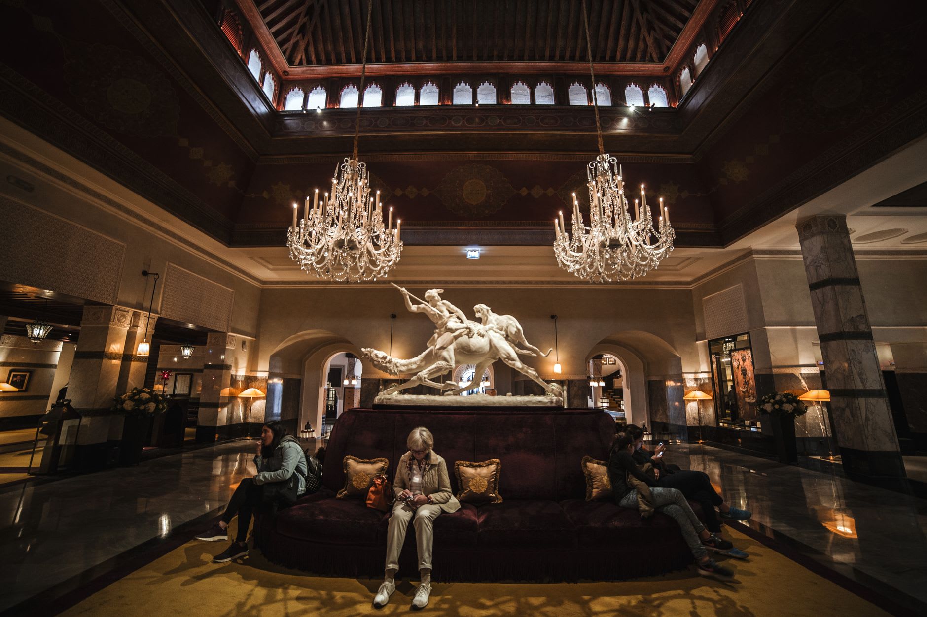Hall d’hôtel de luxe sous deux lustres en cristal, sculpture de marbre et grand canapé de velours rouge