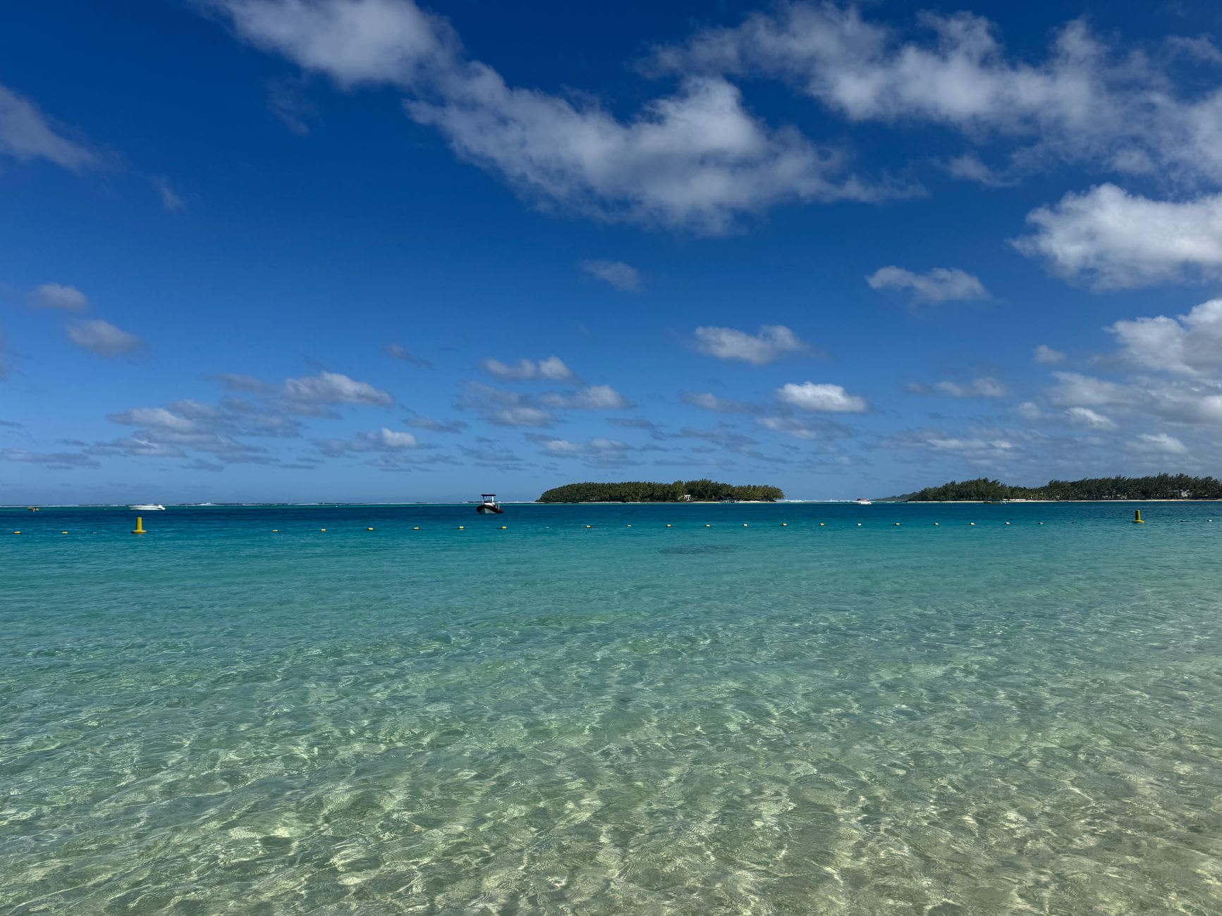 Eau de lagon peu profonde et transparente aux reflets verts, petits îlots boisés à l’horizon sous un ciel bleu parsemé de nuages