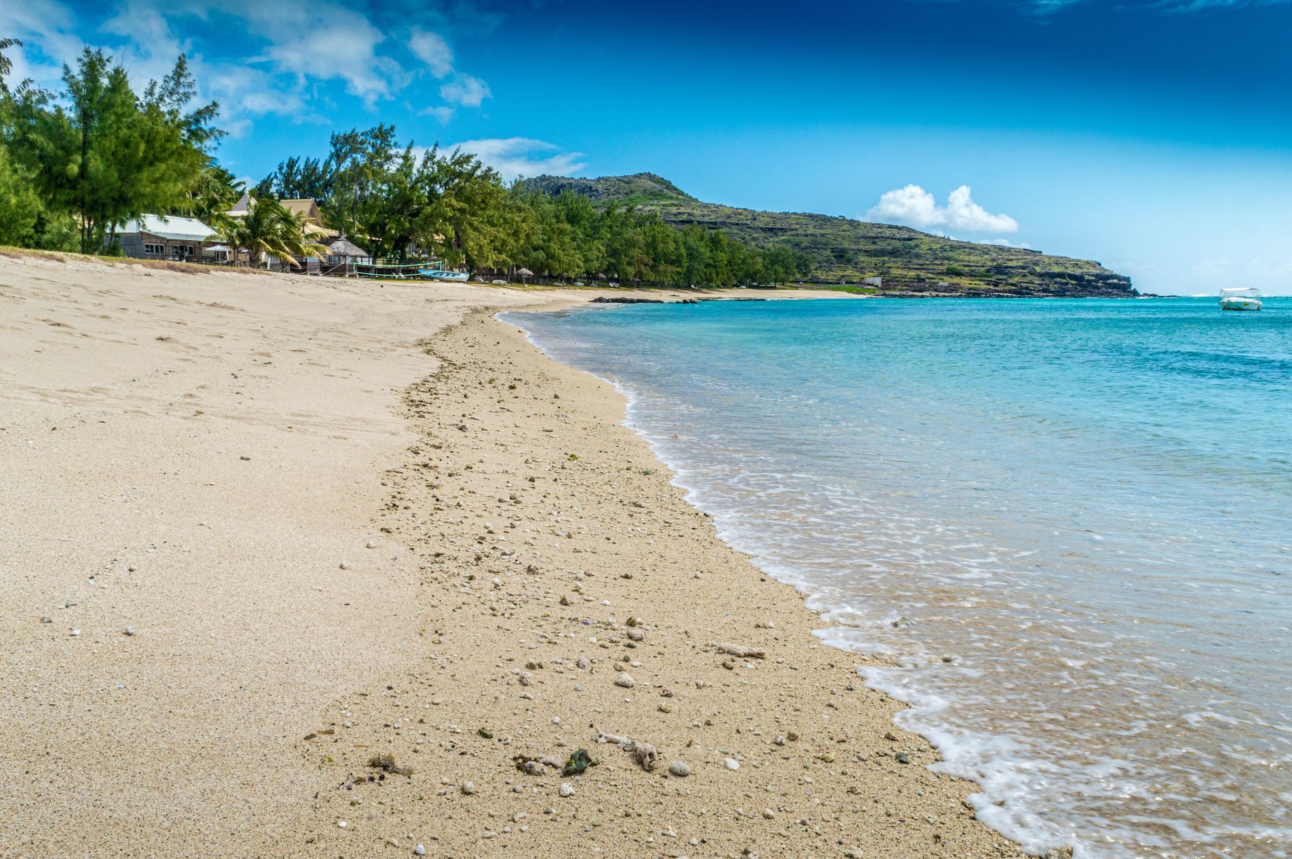 Longue plage de sable clair déserte au bord d’un lagon calme, colline verte et pointe rocheuse fermant la baie