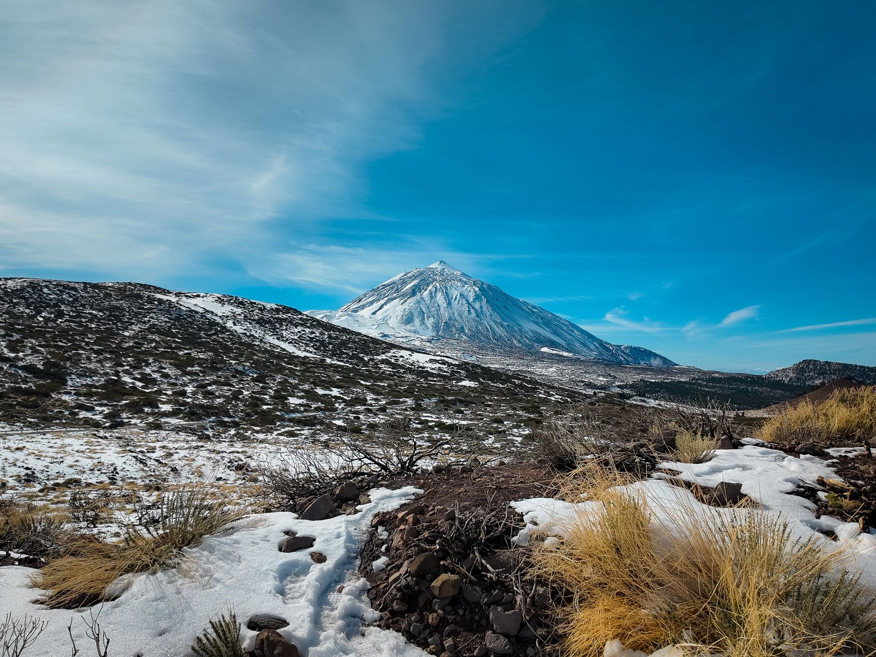 Étendue de neige fraîche et de rochers volcaniques au premier plan, sommet enneigé du volcan au loin sous un ciel bleu