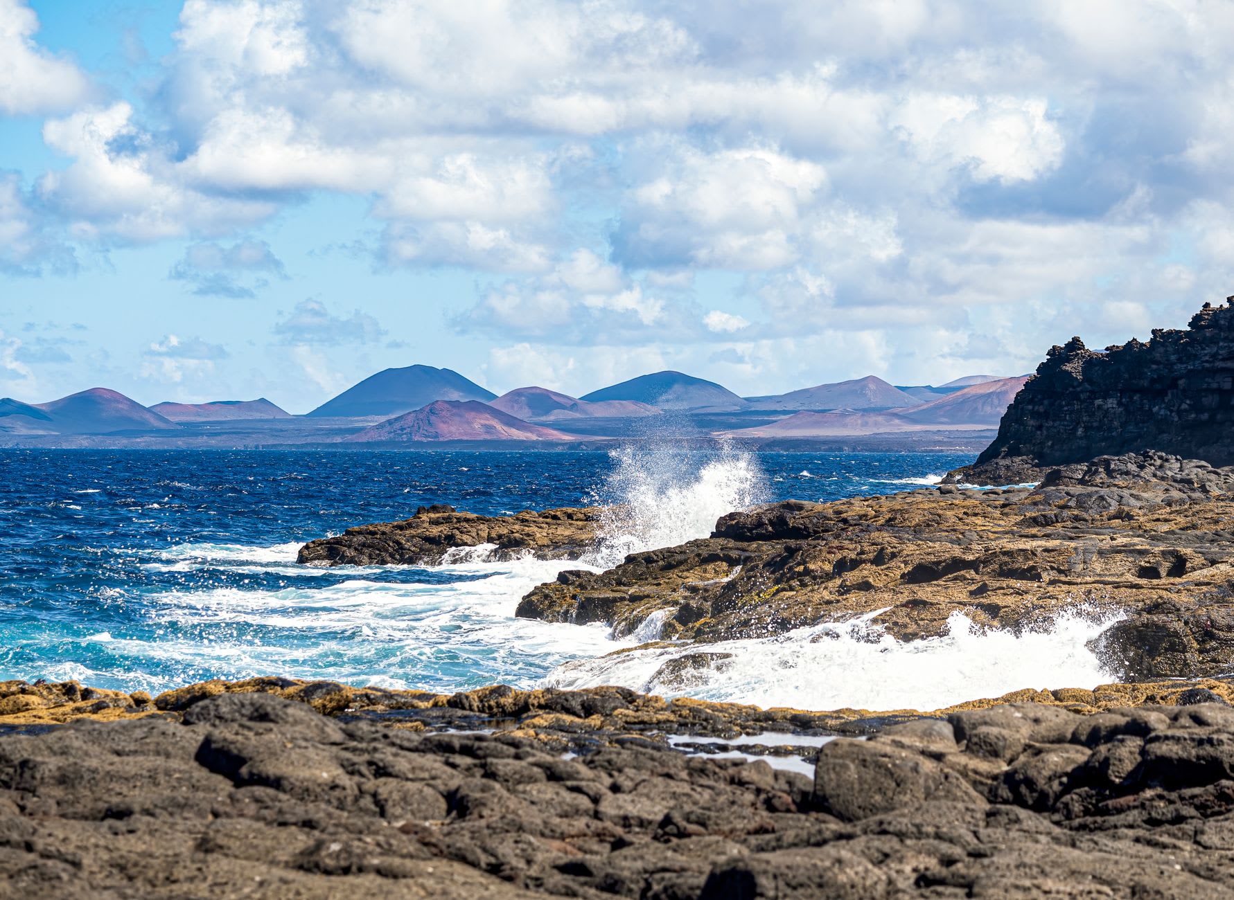Côte rocheuse volcanique battue par les vagues, cônes de volcans bleutés à l’horizon sous un ciel nuageux