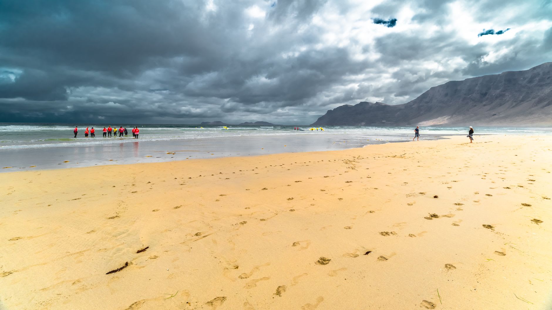 Longue plage de sable dorée battue par les vagues au pied de hautes falaises, ciel d'orage et surfeurs en combinaison au bord de l'eau